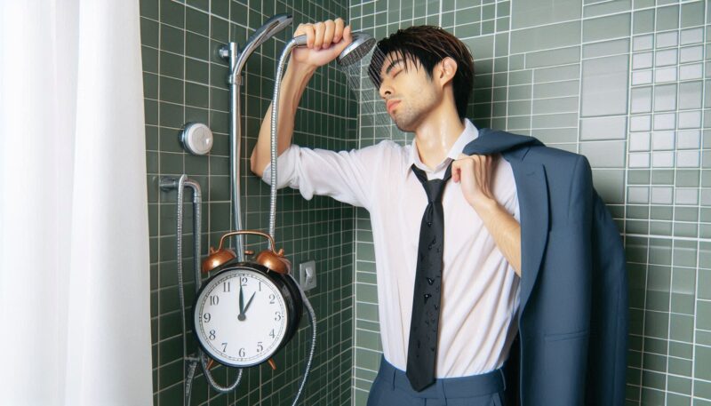 とても眠くて仕事にならなそうなので目を覚まそうとうっかりスーツのままシャワーを浴びている男性。時計は昼休みが終わる13時まであと1分の12時59分を指している。フォトリアリスティックな画像